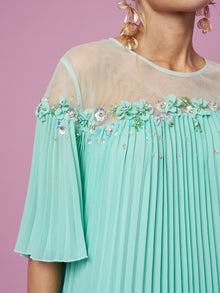A person stands against a pink background wearing the Social Queen Floral Embellishment Pleated Dress in mint green, featuring sheer panels, floral beadwork, sequins, and large gold leaf-shaped earrings.