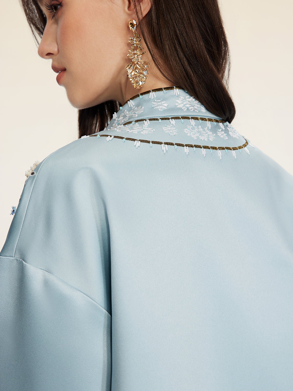 A woman wearing the Social Queen Floral Embroidered Beaded Jacket in light blue is shown from the side and back, highlighting its stand collar, hand-embroidered floral motifs, and her gold dangling earring.