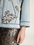 A person wearing the Social Queen Floral Embroidered Beaded Jacket in light blue with hand embroidery, paired with a brown sequin skirt, one hand resting by their side.