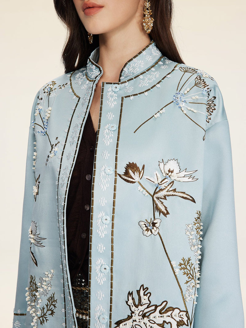 A woman, shown from the shoulders up, wears Social Queen’s Floral Embroidered Beaded Jacket in light blue with hand-embroidered motifs over a black top and gold earrings.