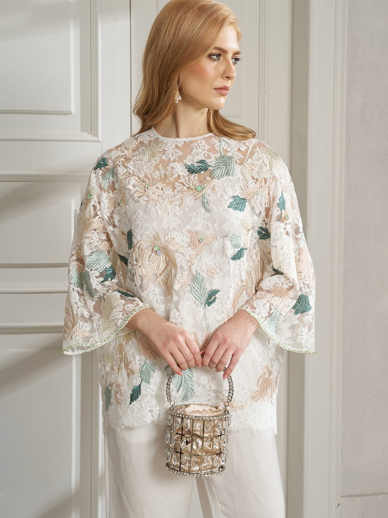 A woman with long blonde hair wears the Social Queen Floral Embroidered Beaded Lace Top and white pants, holding a decorative bag while standing indoors near white paneled doors.
