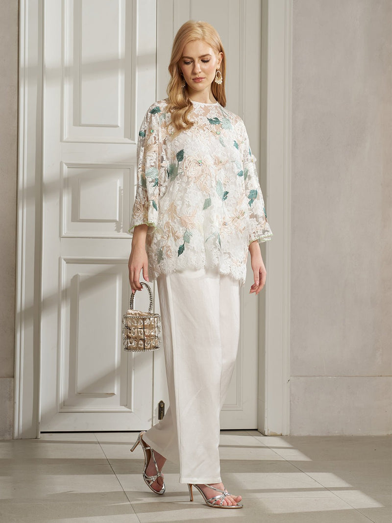 A woman stands indoors in the Social Queen Floral Embroidered Beaded Lace Top, paired with white pants, heeled sandals, and a metallic handbag—an ideal look for festive parties.
