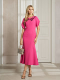 A woman stands indoors in the Social Queen Floral Embroidered Beaded Mermaid Dress, a bright pink, midi-length formal outfit with floral neckline details. She carries a patterned handbag and wears strappy heels.