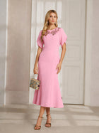 A woman indoors holds a patterned handbag while wearing the Social Queen Floral Embroidered Beaded Mermaid Dress, a light pink, ankle-length formal gown featuring 3D floral appliqué at the neckline.