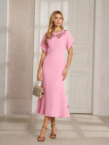 A woman indoors holds a patterned handbag while wearing the Social Queen Floral Embroidered Beaded Mermaid Dress, a light pink, ankle-length formal gown featuring 3D floral appliqué at the neckline.