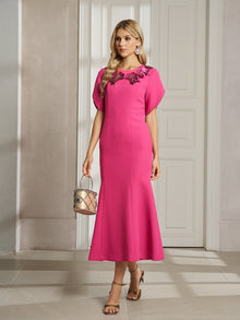 A woman wears the Social Queen Floral Embroidered Beaded Mermaid Dress—rose-red with 3D floral appliqué at the neckline—while holding a metallic handbag and standing indoors on tiled flooring.