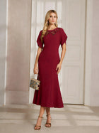 A woman indoors wears the Social Queen Floral Embroidered Beaded Mermaid Dress—calf-length, burgundy with 3D floral details—paired with strappy heels and a metallic handbag.