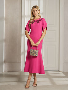 A woman in the Social Queen Floral Embroidered Beaded Mermaid Dress stands indoors on a tiled floor, holding a small patterned handbag.