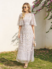 A woman stands outdoors in sunlight wearing the Social Queen Floral Embroidered Beaded Tulle Dress with puff sleeves, sunglasses, and holding a small purse, surrounded by flowers and greenery.
