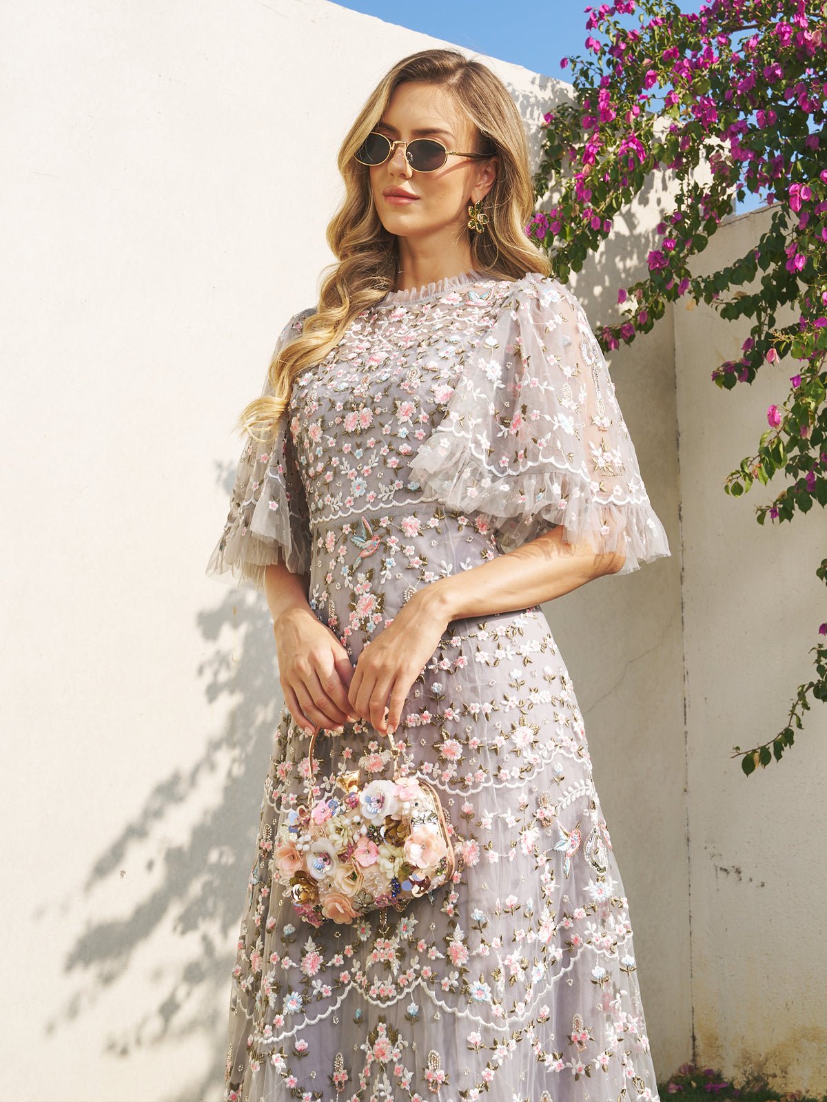 A woman in a Social Queen Floral Embroidered Beaded Tulle Dress with puff sleeves stands outdoors, holding a matching floral purse and wearing sunglasses, with bougainvillea blooming in the background.