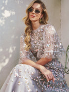 A woman in the Social Queen Floral Embroidered Beaded Tulle Dress and sunglasses sits on a chair against a light wall, with sunlight casting soft shadows.