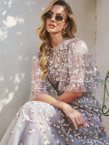 A woman in the Social Queen Floral Embroidered Beaded Tulle Dress and sunglasses sits on a chair against a light wall, with sunlight casting soft shadows.