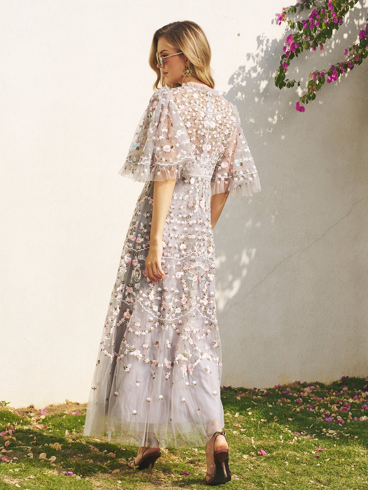 A woman stands outdoors near a white wall and flowering vine, wearing the Social Queen Floral Embroidered Beaded Tulle Dress with puff sleeves and heels, facing away from the camera.