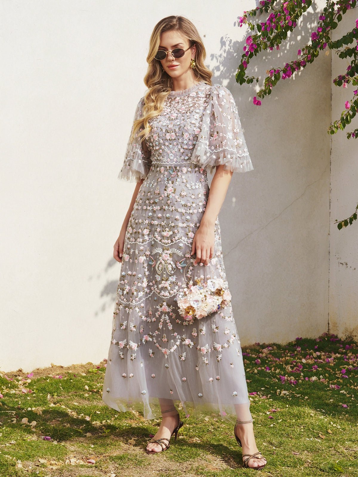 A woman wears the Social Queen Floral Embroidered Beaded Tulle Dress—light gray with puff sleeves—holding a matching purse and standing on grass near a white wall beneath pink flowers.