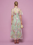 A woman, facing away against a pink background, wears the Social Queen Floral Embroidered Beaded Tulle Gown with sheer short sleeves, paired with pink heels and a woven bag.
