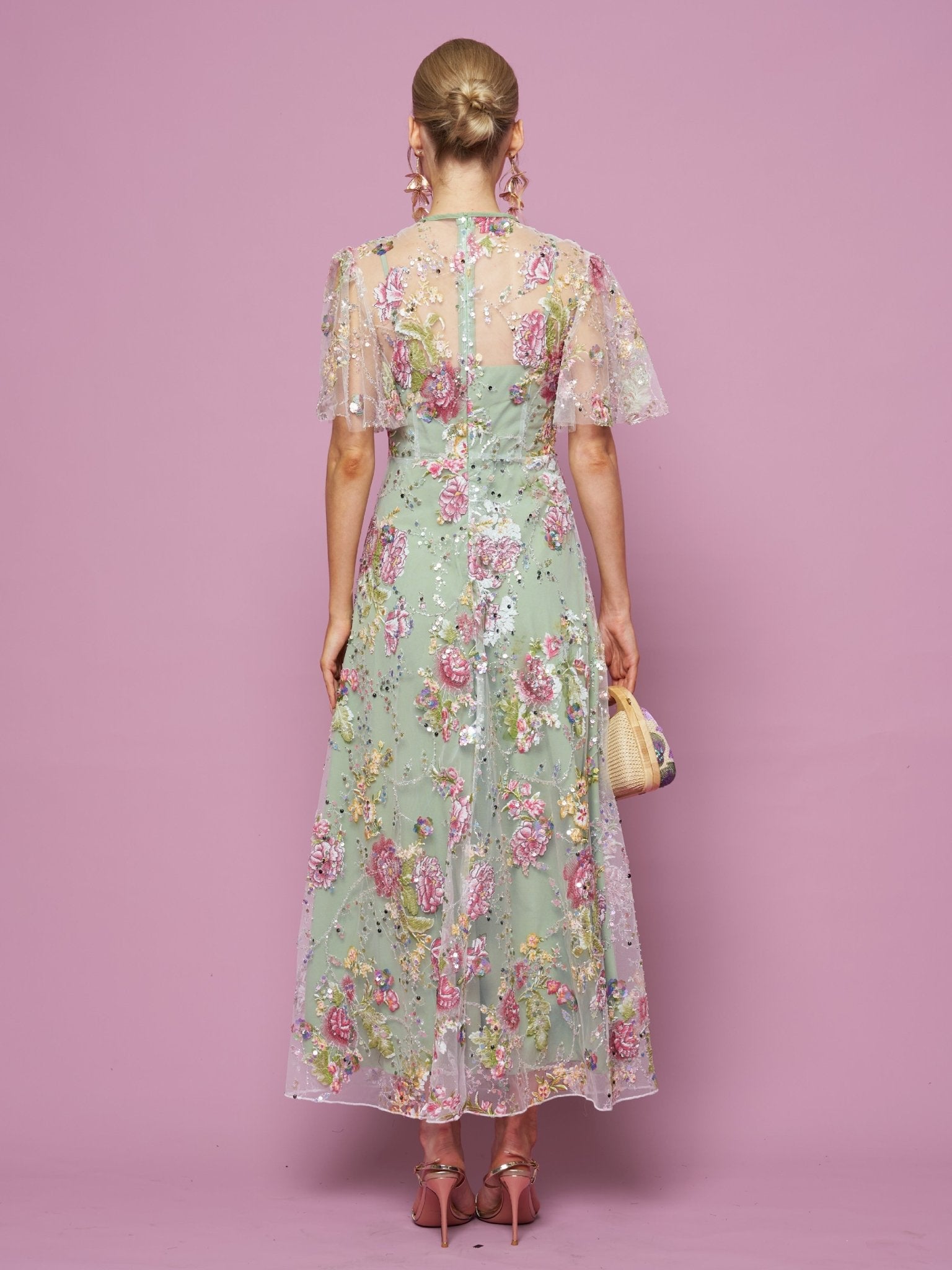 A woman, facing away against a pink background, wears the Social Queen Floral Embroidered Beaded Tulle Gown with sheer short sleeves, paired with pink heels and a woven bag.