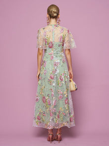 A woman, facing away against a pink background, wears the Social Queen Floral Embroidered Beaded Tulle Gown with sheer short sleeves, paired with pink heels and a woven bag.