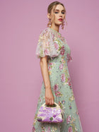 A woman in Social Queen's Floral Embroidered Beaded Tulle Gown holds a small purple handbag, posed against a solid pink background.