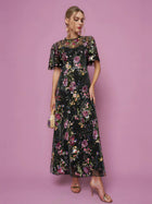 A woman wearing the Social Queen Floral Embroidered Beaded Tulle Gown with short sleeves holds a small handbag while posing against a pink background.