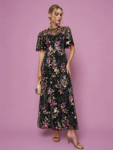 A woman wearing the Social Queen Floral Embroidered Beaded Tulle Gown with short sleeves holds a small handbag while posing against a pink background.