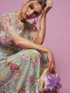 A woman in a Social Queen Floral Embroidered Beaded Tulle Gown sits against a pink background, resting her head on her hand and holding a small purse with a purple floral pattern.
