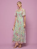 A woman poses against a pink backdrop in the Social Queen Floral Embroidered Beaded Tulle Gown with short sheer sleeves, holding a small purse and wearing heeled sandals.