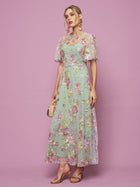 A woman poses against a pink backdrop in the Social Queen Floral Embroidered Beaded Tulle Gown with short sheer sleeves, holding a small purse and wearing heeled sandals.