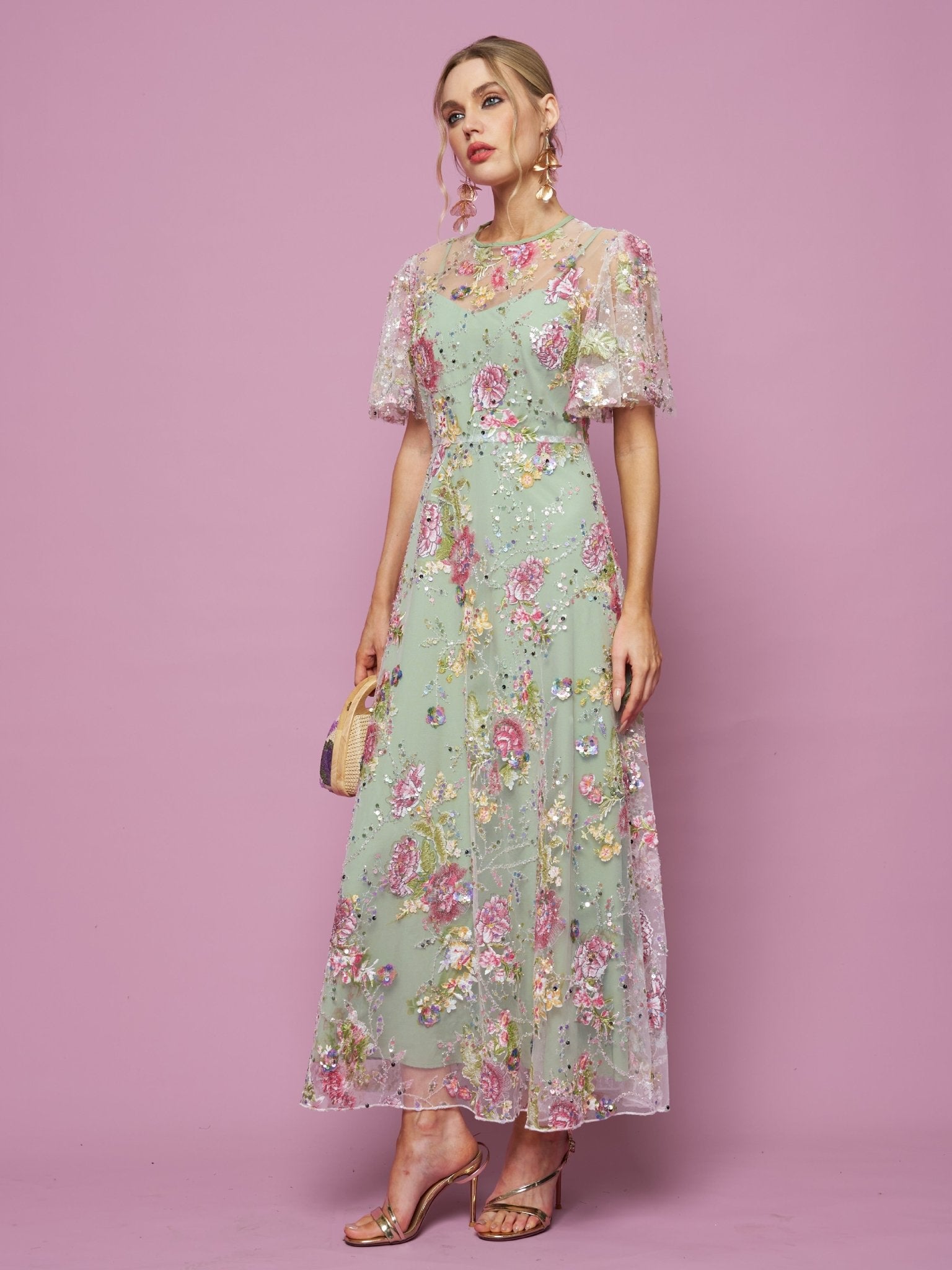 A woman poses against a pink backdrop in the Social Queen Floral Embroidered Beaded Tulle Gown with short sheer sleeves, holding a small purse and wearing heeled sandals.