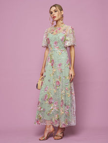 A woman poses against a pink backdrop in the Social Queen Floral Embroidered Beaded Tulle Gown with short sheer sleeves, holding a small purse and wearing heeled sandals.