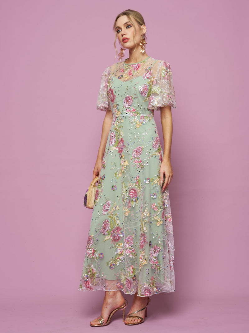 A woman poses against a pink backdrop in the Social Queen Floral Embroidered Beaded Tulle Gown with short sheer sleeves, holding a small purse and wearing heeled sandals.