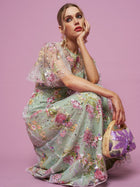 A person in the Social Queen Floral Embroidered Beaded Tulle Gown sits on the floor against a pink backdrop, holding a round bag with purple patterns and resting their chin on their hand.