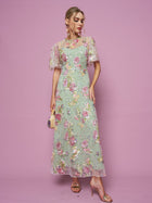Against a pink background, a person wears the Social Queen Floral Embroidered Beaded Tulle Gown, paired with a small purse and gold high-heeled sandals.