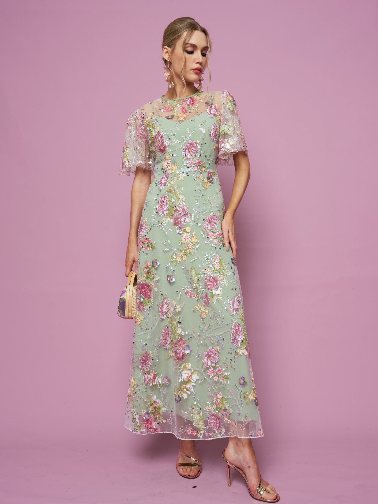 Against a pink background, a person wears the Social Queen Floral Embroidered Beaded Tulle Gown, paired with a small purse and gold high-heeled sandals.