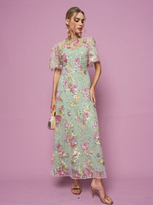 Against a pink background, a person wears the Social Queen Floral Embroidered Beaded Tulle Gown, paired with a small purse and gold high-heeled sandals.