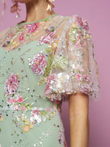 Close-up of a woman in the Social Queen Floral Embroidered Beaded Tulle Gown, featuring pastel green tulle sleeves and colorful embellishments, set against a pink background.