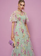 A woman stands against a pink background in a mint green Floral Embroidered Beaded Tulle Gown by Social Queen, featuring sheer sleeves, and holds a small purple and yellow handbag.