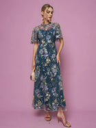 A woman in the Social Queen Floral Embroidered Beaded Tulle Gown and gold heels holds a small beige clutch, posing against a plain pink background.