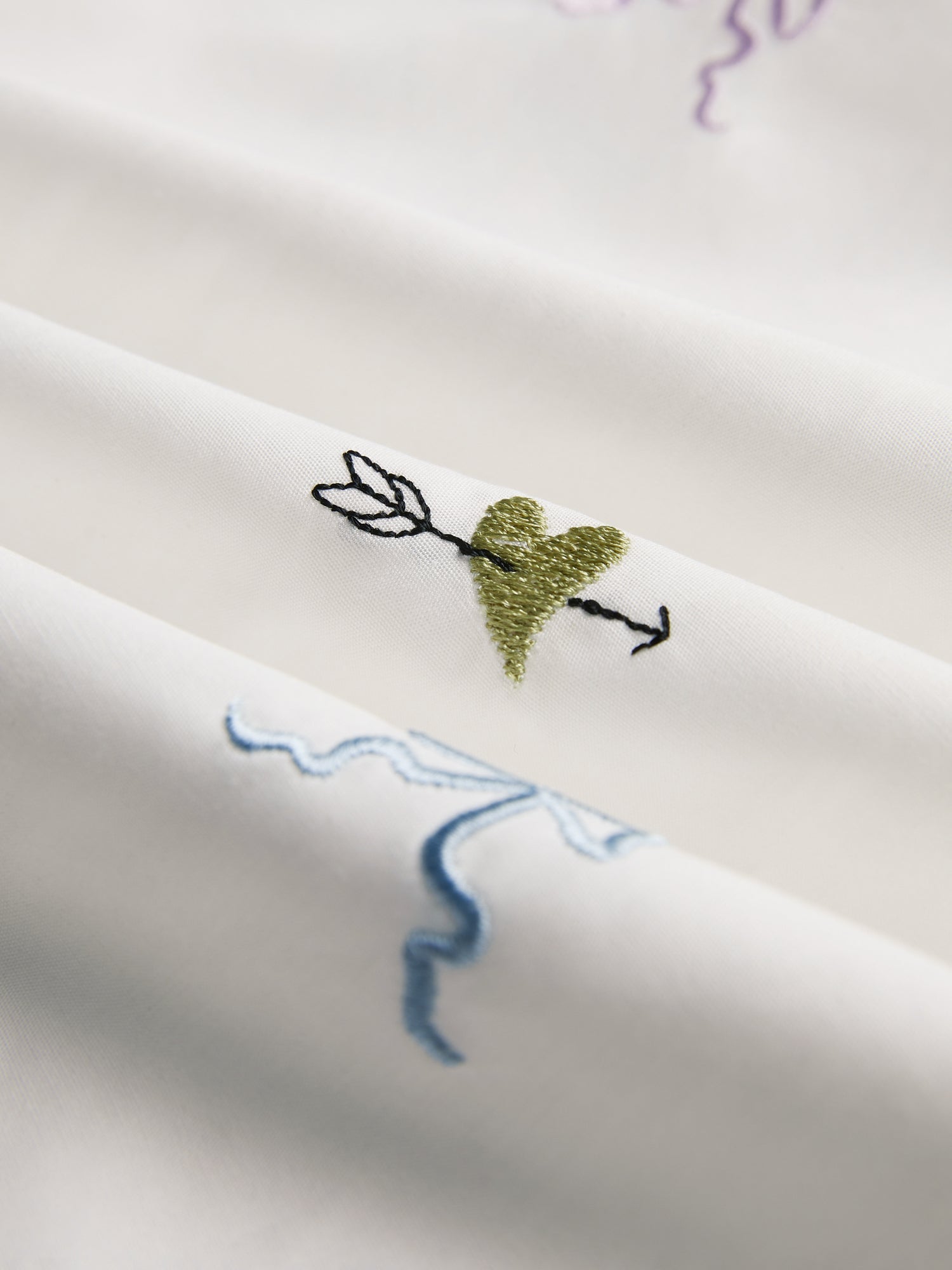 Close-up of the Floral Embroidered Cotton Blouse by Social Queen in white, showcasing a green heart with an arrow and light blue and purple abstract lines.