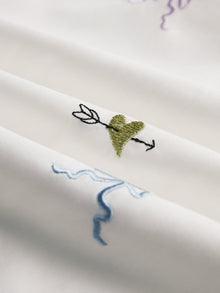 Close-up of the Floral Embroidered Cotton Blouse by Social Queen in white, showcasing a green heart with an arrow and light blue and purple abstract lines.