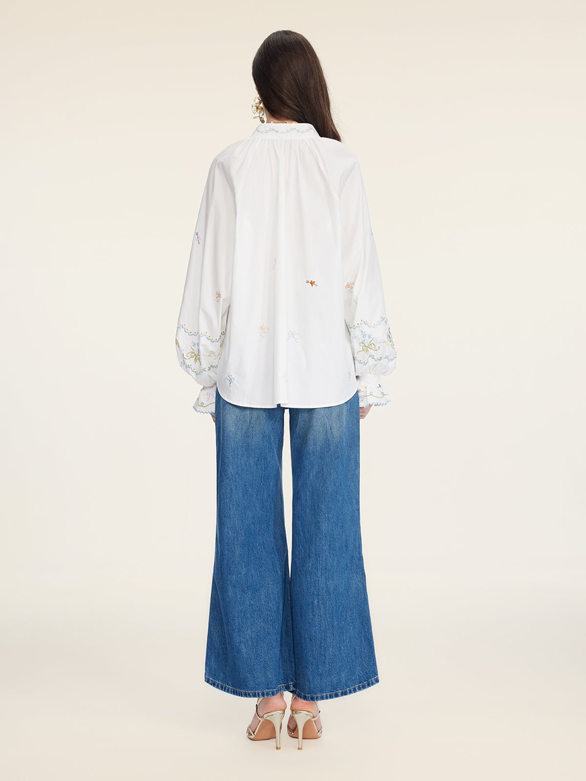 A person with long dark hair faces away, wearing Social Queen’s Floral Embroidered Cotton Blouse with long sleeves, wide-leg blue jeans, and heeled sandals.