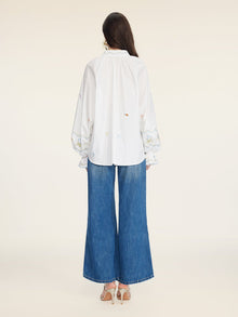 A person with long dark hair faces away, wearing Social Queen’s Floral Embroidered Cotton Blouse with long sleeves, wide-leg blue jeans, and heeled sandals.