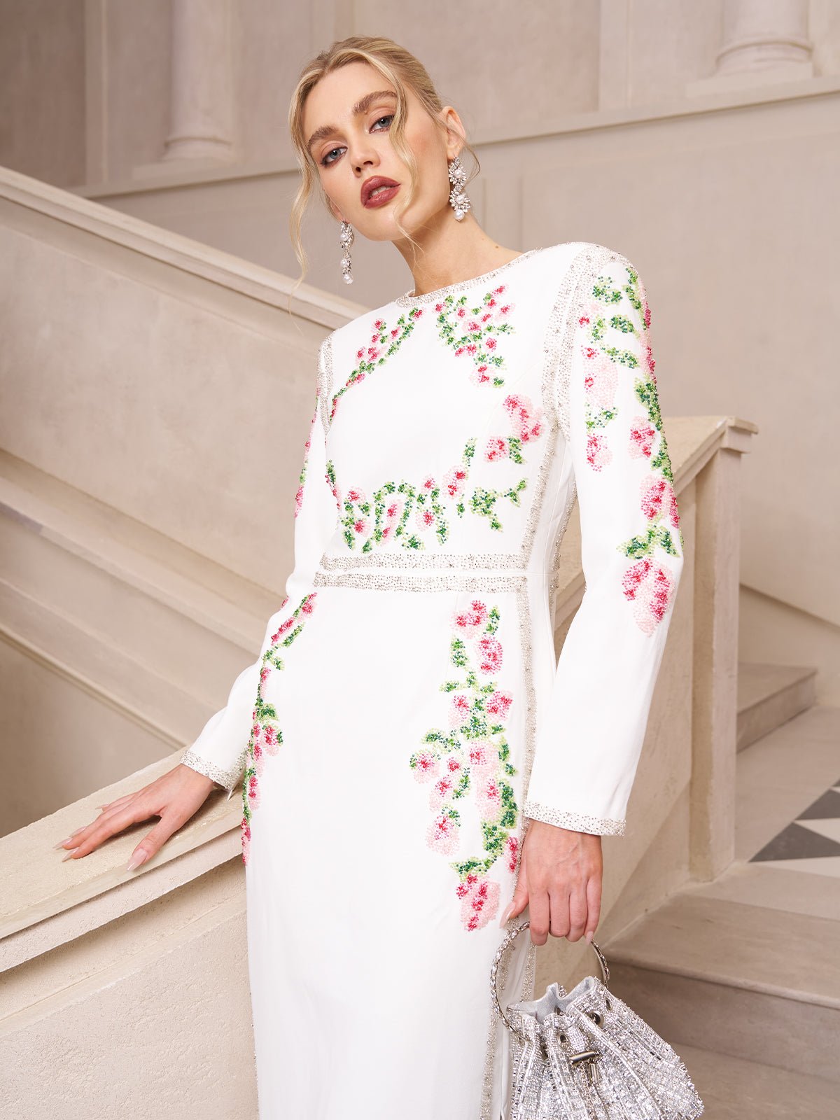 A woman wears the Social Queen Floral Embroidered Crystal Dress, featuring pink and green floral details, paired with a silver handbag and statement earrings—an elegant choice for evening occasions.