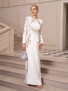 A woman wears the Social Queen Floral Embroidered Crystal Dress—gown-length white with pink and green embroidery—paired with silver heels, earrings, and a handbag, ideal for an elegant evening event.