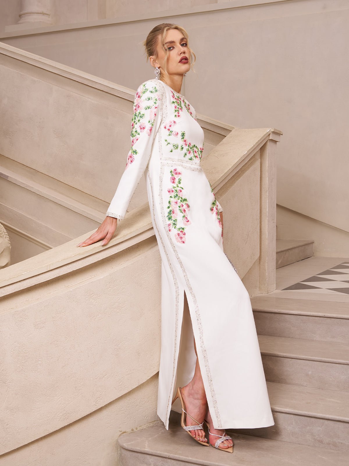 A woman wears the Social Queen Floral Embroidered Crystal Dress, featuring pink floral embroidery, as she stands on a staircase with one arm resting on the railing.