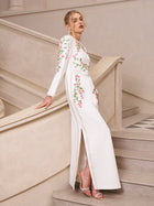 A woman wears the Social Queen Floral Embroidered Crystal Dress, featuring pink floral embroidery, as she stands on a staircase with one arm resting on the railing.