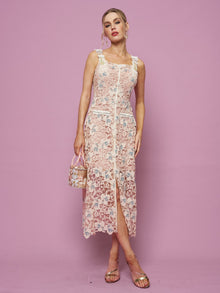 A woman stands against a pink background in the Social Queen Floral Embroidered Cutout Lace Dress, accessorized with a small handbag and open-toe sandals for an ideal semi-formal look.