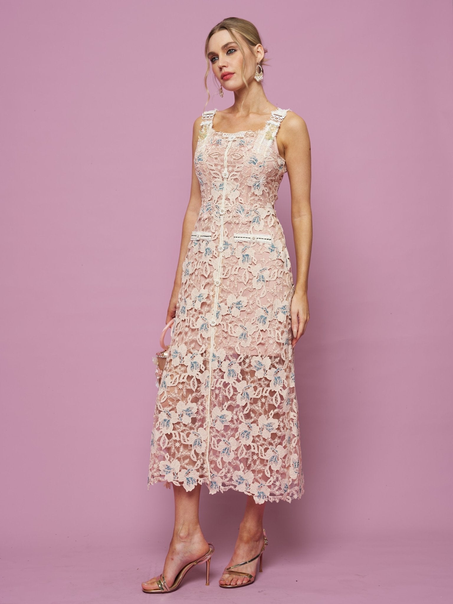 A woman poses against a pink background in the Social Queen Floral Embroidered Cutout Lace Dress—a sleeveless, knee-length, pale pink semi-formal dress with delicate floral embroidery—paired with strappy high-heeled sandals.