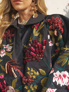 A woman wears Social Queen's Floral Embroidered Floral Puff Sleeve Jacket, featuring vibrant hand-embroidered florals, paired with gold leaf earrings.