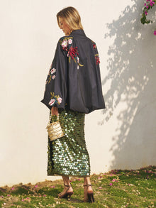 A woman stands on grass in profile, wearing the Social Queen Floral Embroidered Puff Sleeve Jacket, a green sequin skirt, heeled sandals, and holding a beige woven handbag.