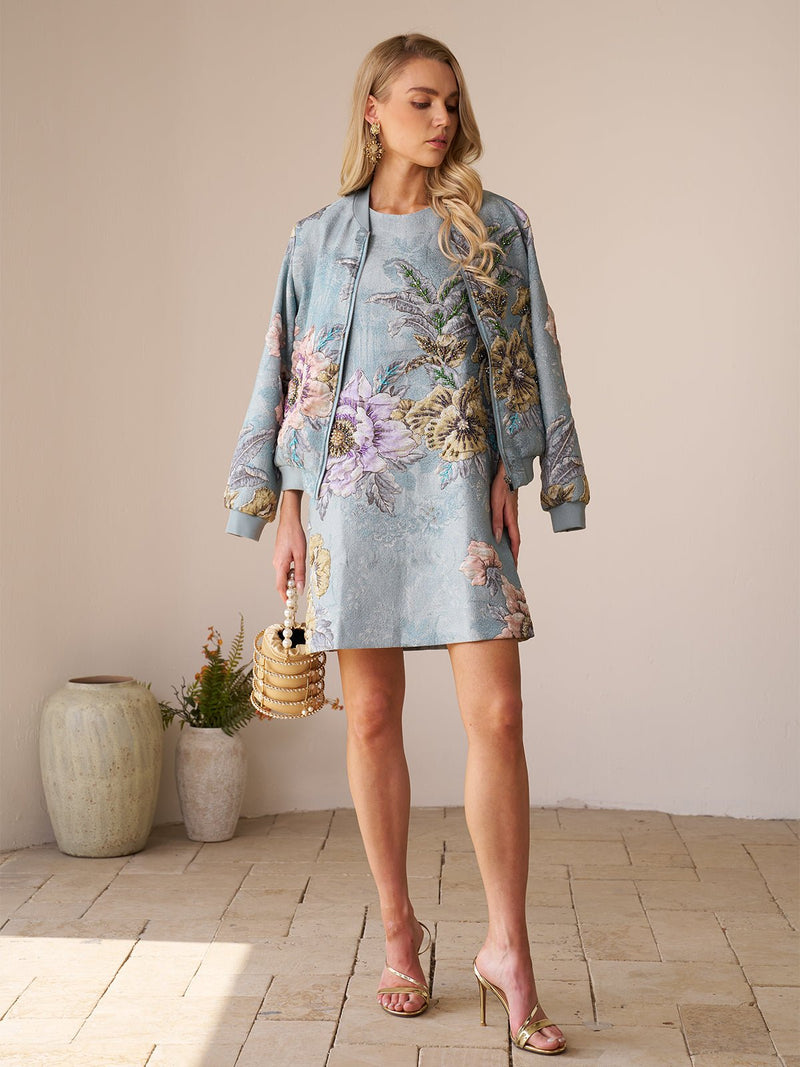 Indoors, a woman wears a light blue floral dress and the Floral Embroidered Jacquard Jacket by Social Queen, holding a small handbag and wearing gold sandals. Two vases with plants are in the background.